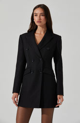 The woman is wearing a tailored black blazer dress with a double-breasted design, featuring six buttons on the front. The blazer has a notched lapel collar, structured shoulders, and long sleeves. The dress length is above the knees, revealing sheer black tights.